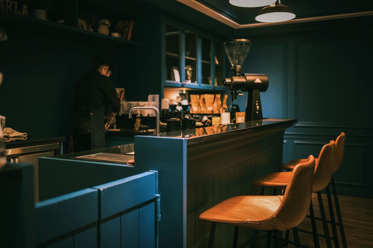 Dunkler Innenraum des Cafés: Bar mit Maschine, Mühle und einer Reihe kupferner Hocker.
