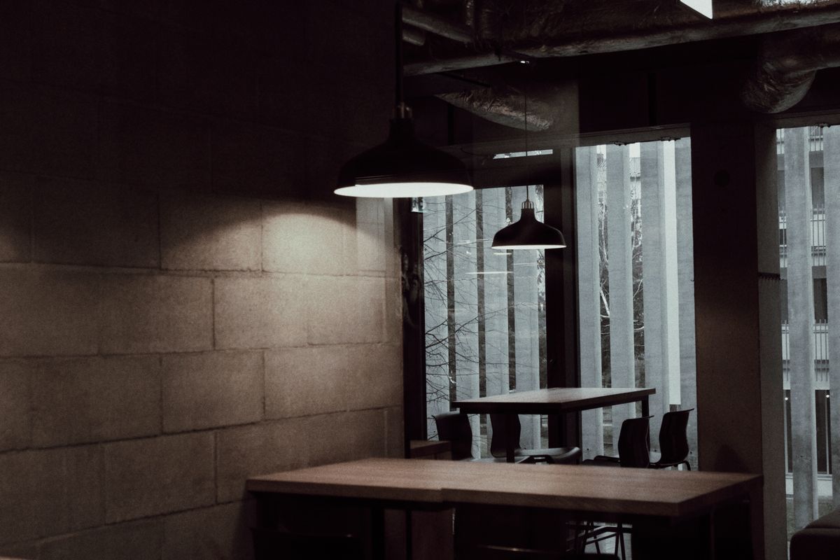 Gastraum in dunklen Tönen, Betonwand und zwei Lampen über dem Tisch am Fenster.