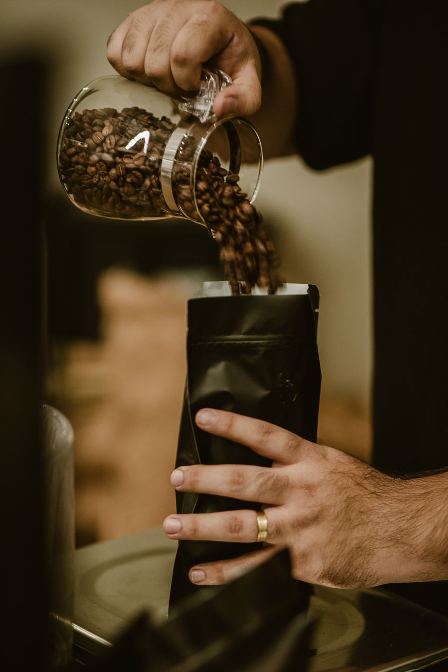 Eine Hand füllt geröstete Bohnen aus einem Glas in eine schwarze Kaffeetüte.