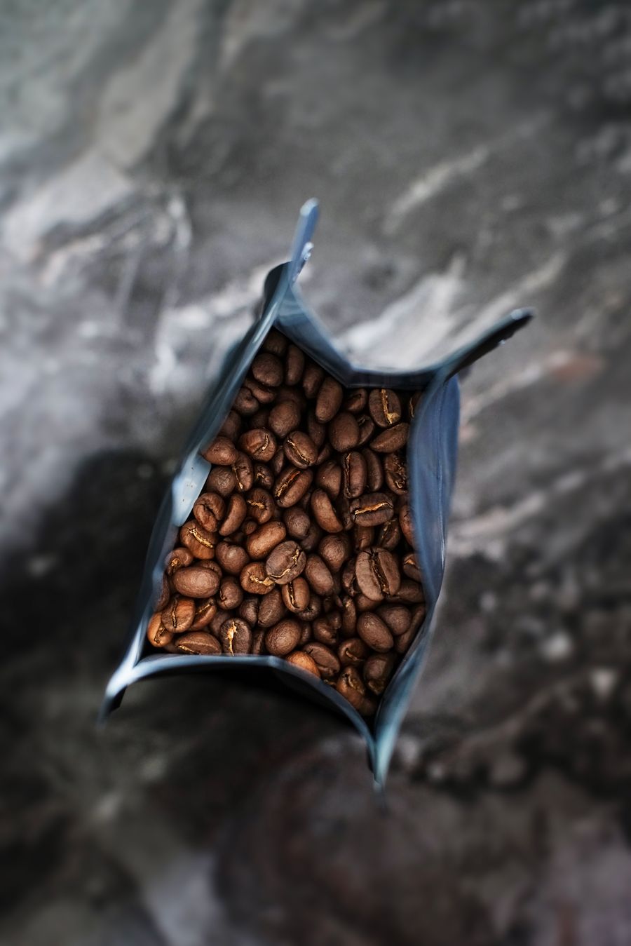 Offene Tüte voller gerösteter Kaffeebohnen auf dunkler Steinplatte.