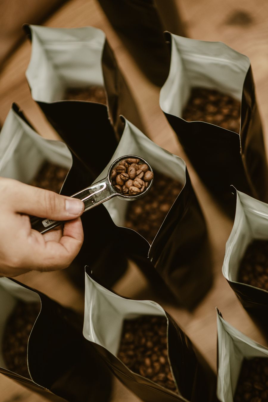 Eine Hand mit einem Messbecher voller Bohnen über einer Reihe offener Kaffeetüten.